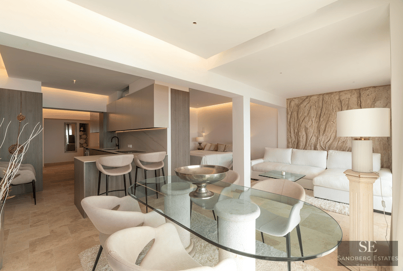 Modernt vardagsrum med matbord i glas, krämvit soffa och integrerat kök i neutrala toner och sten.