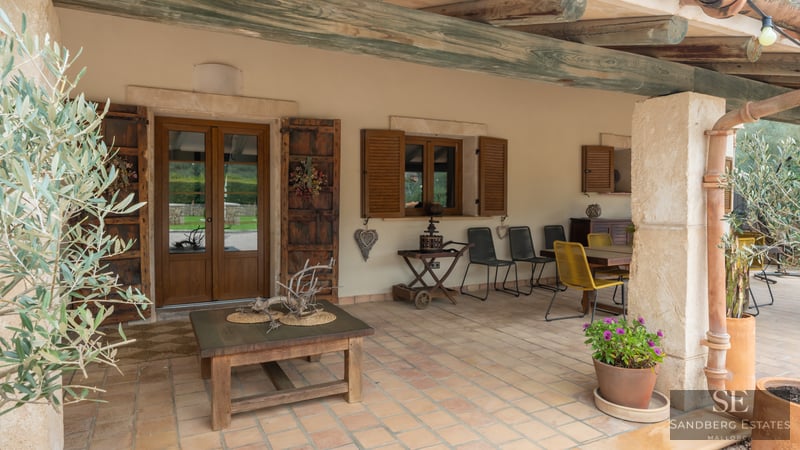 Porche cubierto con baldosas de terracota, vigas de madera rústicas, muebles de comedor y paredes de piedra clara.