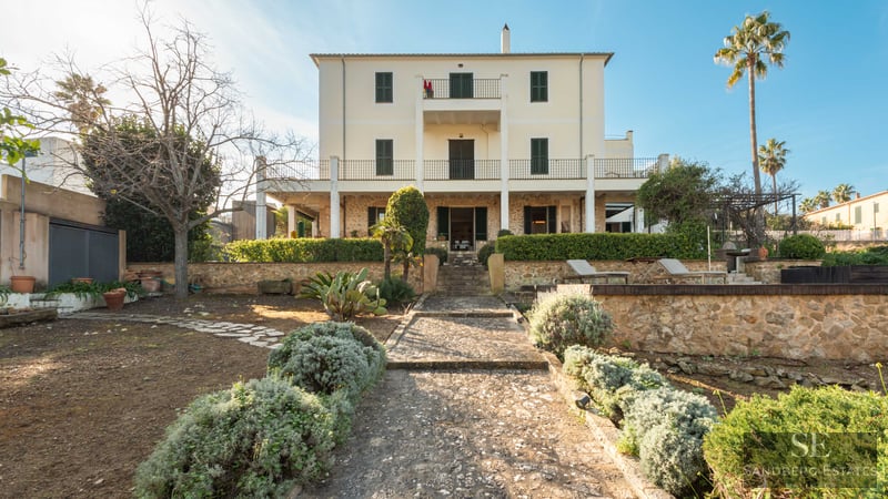 Rear view of a cream three-story villa with stone walls, green shutters, and a stone path through a garden.