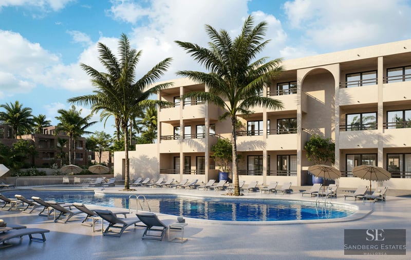 Grande piscine bleue avec chaises longues, palmiers et bâtiment méditerranéen moderne de trois étages sous un ciel clair.