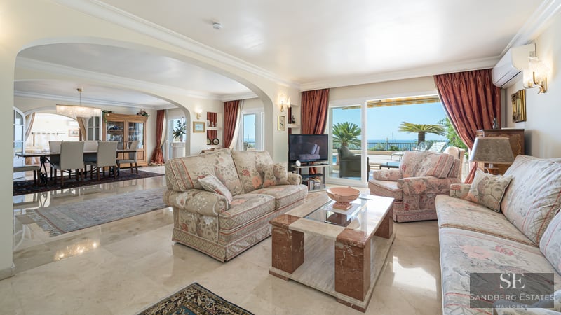 Bright living room with floral sofas, marble coffee table, and large glass doors opening to a terrace with sea view.