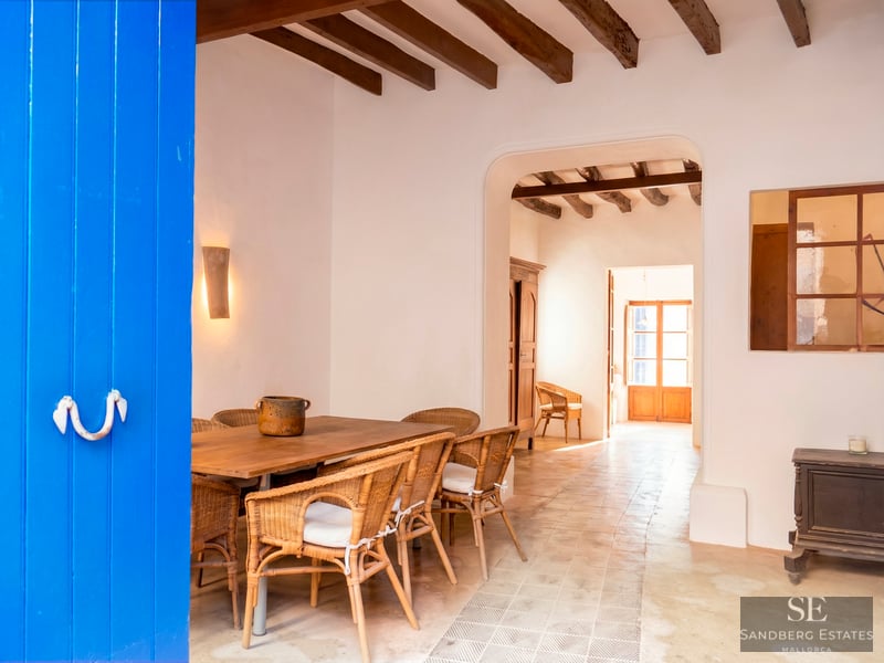 Dining area featuring a wooden table, wicker chairs, exposed ceiling beams, and a striking blue door in the foreground.