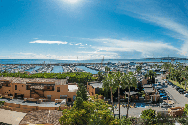 Elevated view of a Mediterranean port with yachts, terracotta buildings, and palm trees under a bright blue sky.