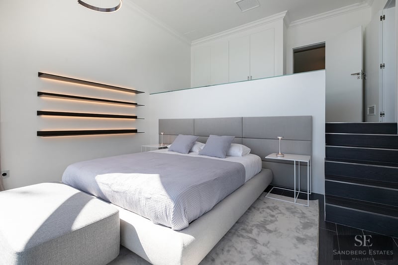 Modern bedroom featuring a gray bed, integrated LED wall shelving, and minimalist white interior.