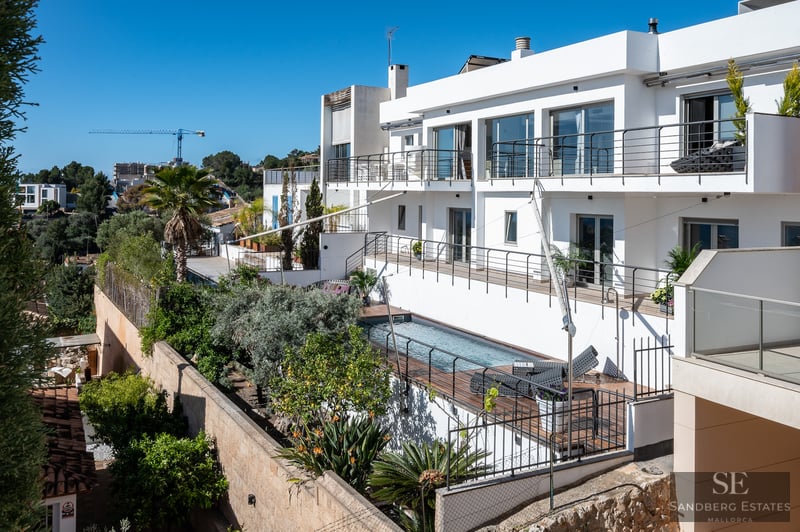 Villa moderna blanca de varios niveles con piscina rectangular, cubierta de madera y exuberante jardín mediterráneo.