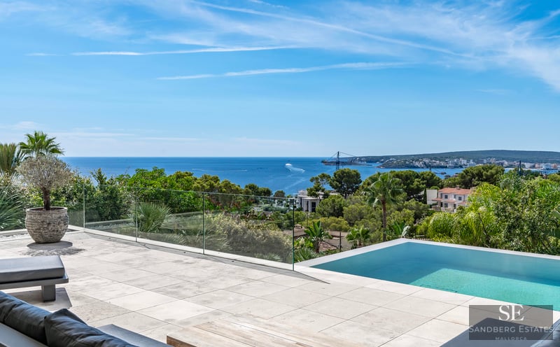 Modern stone terrace featuring an infinity pool with a glass railing overlooking the blue sea and lush greenery.
