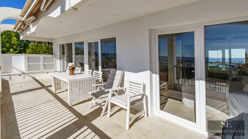 Sunlit terrace with white outdoor dining set and large glass sliding doors reflecting the coastline.