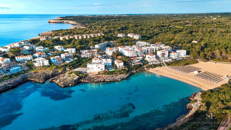 Vue aérienne par drone d'une baie turquoise avec une plage de sable entourée de bâtiments blancs et d'une forêt verte.