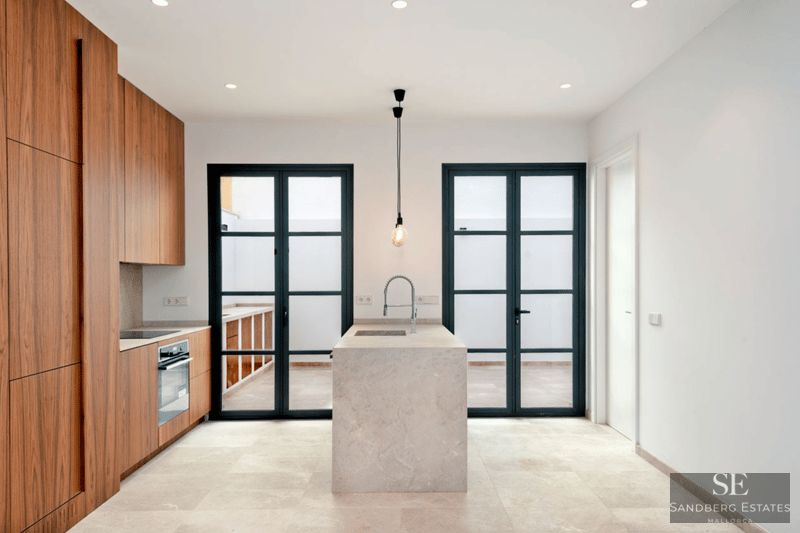 Minimalistische Küche mit zentraler Insel aus beigem Stein, Walnussholzschränken und großen schwarzen Glastüren.