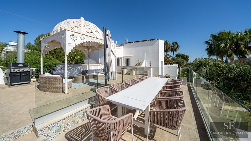 Terraza amplia con mesa de comedor blanca, sillas de mimbre, barbacoa y un cenador blanco ornamentado bajo el sol.