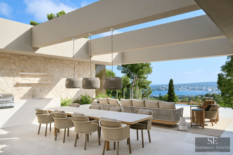 Terraza moderna con una gran mesa de comedor, cocina exterior y sofás con vistas al mar Mediterráneo y la costa.