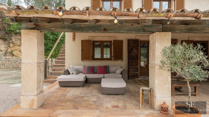 Terraza cubierta con vigas de madera, columnas de piedra, sofá cómodo y un olivo en maceta.