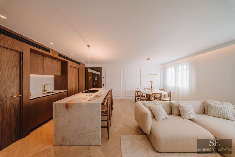 Modern open-plan room with a marble kitchen island, wood cabinets, herringbone floors, and a large cream sectional sofa.