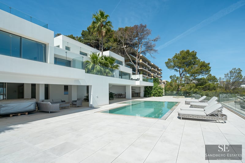 Modern white villa with a rectangular swimming pool, grey sun loungers, and glass railings under a clear blue sky.
