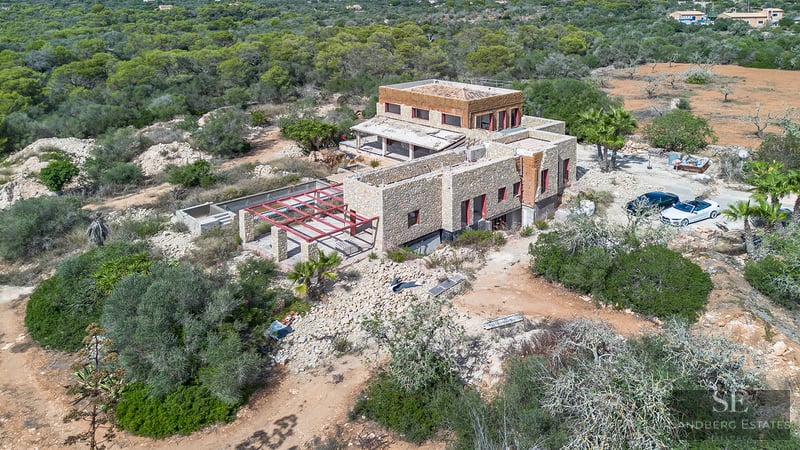 Vista aérea de una gran villa de piedra en construcción rodeada de densa vegetación mediterránea.