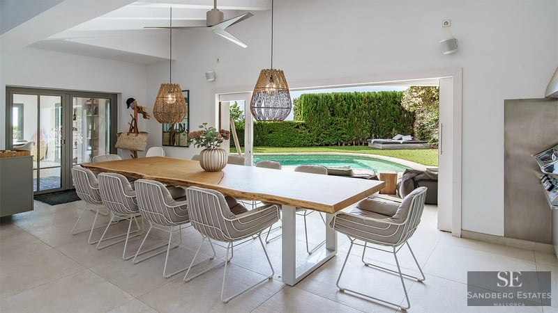 Elegante comedor con gran mesa de madera y lámparas de mimbre, abierto a una terraza con piscina y jardín.
