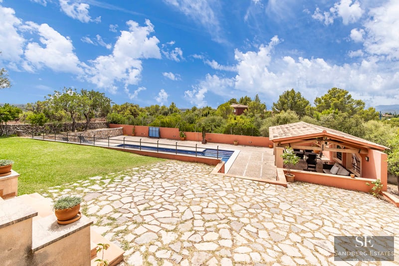 Gran patio de piedra con piscina, césped verde y una zona de estar cubierta con vigas de madera y techo de tejas.