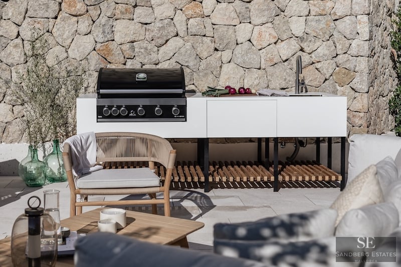 Modern white outdoor kitchen with gas grill and sink against a stone wall with designer patio furniture.