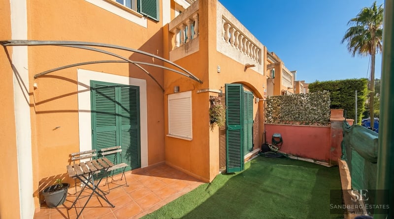 Small private garden with artificial grass, terracotta tiles, green shutters, and a wooden bistro set under a sunny sky.