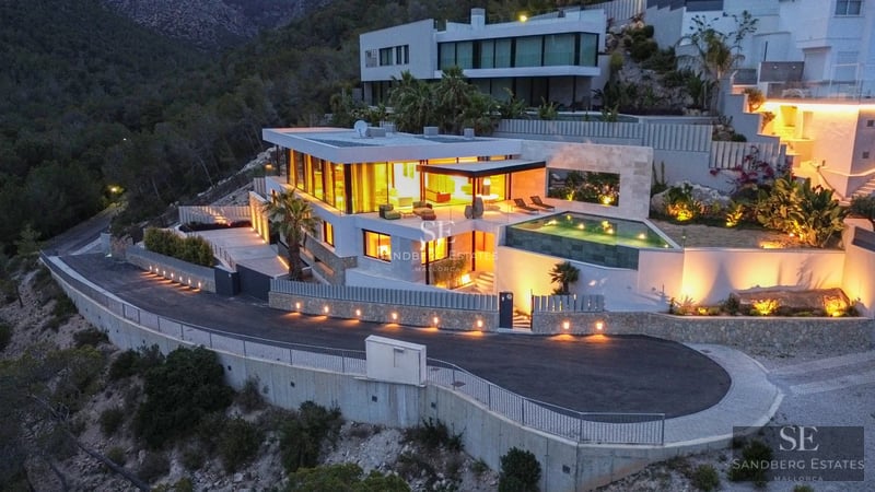 View of a luxurious villa with infinity pool, landscaped gardens, and modern architecture. Stone facade and large windows.
