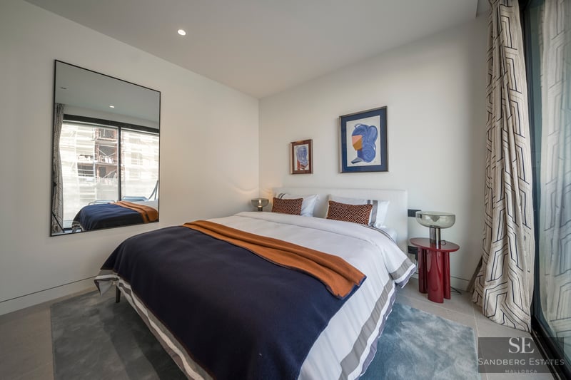 Dormitorio moderno con cama grande, ropa de cama azul y naranja, mesa roja y un gran espejo que refleja una ventana.