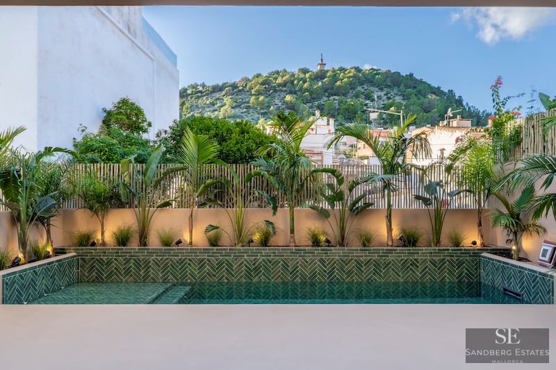 Piscine moderne avec carreaux verts en chevrons entourée de plantes tropicales et vue sur une colline sous un ciel bleu.