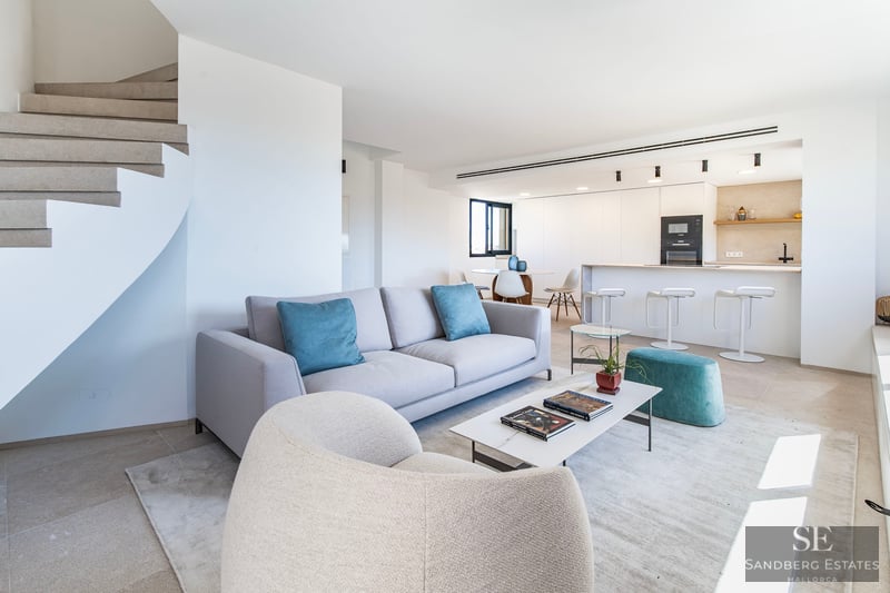 Salón moderno con sofá gris, cojines verde azulado, mesa de mármol y escalera de caracol junto a cocina blanca.