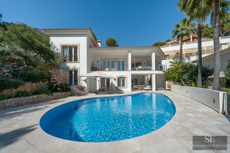 Grande piscine ovale bleu azur devant une villa blanche à deux étages avec terrasse en pierre et palmiers.