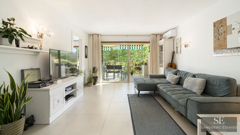 Salon moderne avec canapé bleu, meuble TV blanc et baies vitrées ouvrant sur une terrasse verdoyante.