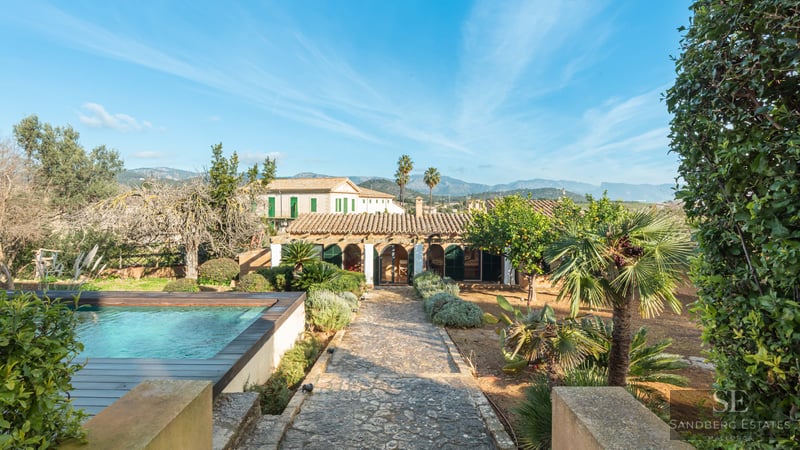 Outdoor swimming pool and stone path leading to a Mediterranean villa with mountain views under a blue sky.