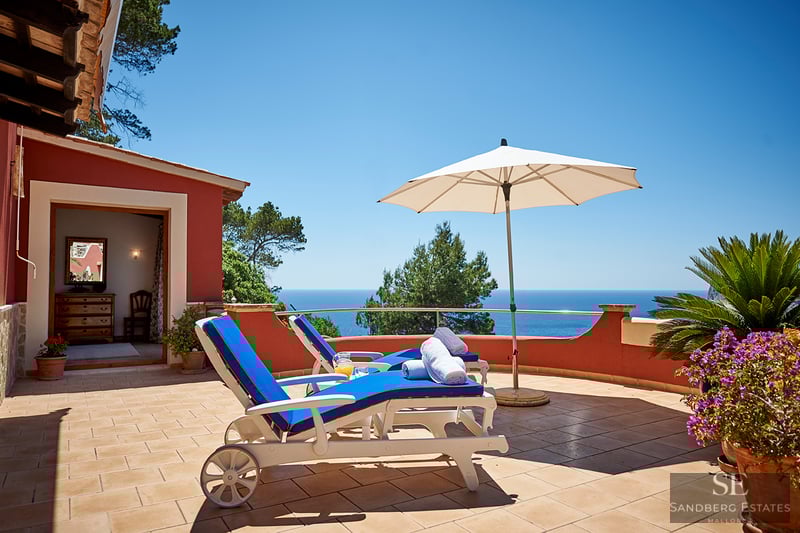 Två blå solstolar på en terrakottaterrass med utsikt över ett klarblått hav under en molnfri himmel.