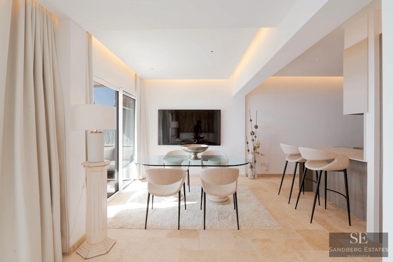 Minimalistisk matsal med glasbord, cremefärgade stolar, vit matta och frukostbar i bakgrunden.