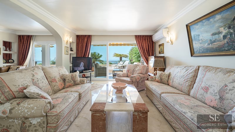 Spacious living room with floral sofas, marble table, and large glass doors opening to a terrace overlooking the sea.