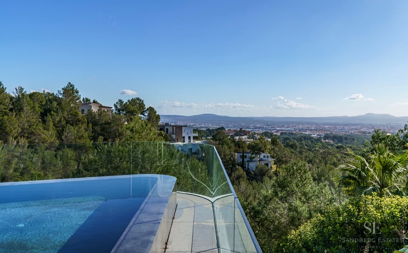 An infinity-edge pool with glass railing overlooking a lush forested valley and distant city.
