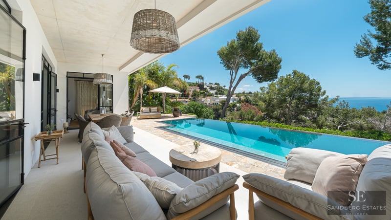 Covered terrace with grey lounge seating overlooking a turquoise infinity pool and the Mediterranean sea.