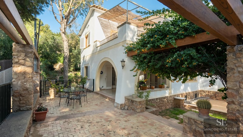 Sunny tiled patio with a flowering wooden pergola and white villa facade featuring arched entrance.