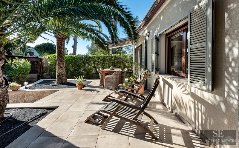 Sunny tiled patio with wooden lounge chairs, a dining set, and a large palm tree against a beige house wall with shutters.