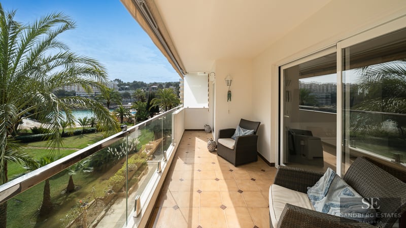 Sunlit balcony with wicker chairs, glass railing, and a scenic view of lush palm trees and a garden.