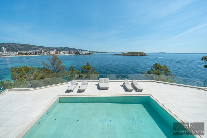 Modern swimming pool and sun loungers on a white terrace overlooking a turquoise bay and distant coastline.