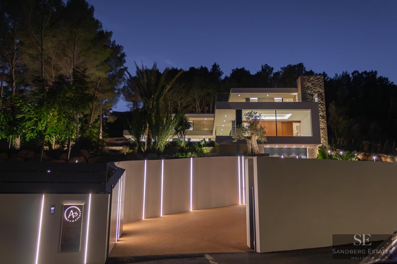 Modern white luxury villa at night with illuminated LED driveway, glass walls, and lush palm trees against a dark sky.