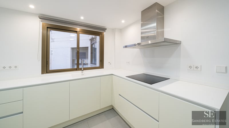 Minimalist white kitchen featuring L-shaped counter, induction stove, stainless steel extractor, and window.