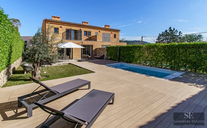 Modern outdoor pool and wooden deck with sun loungers next to a terracotta villa under a clear blue sky.