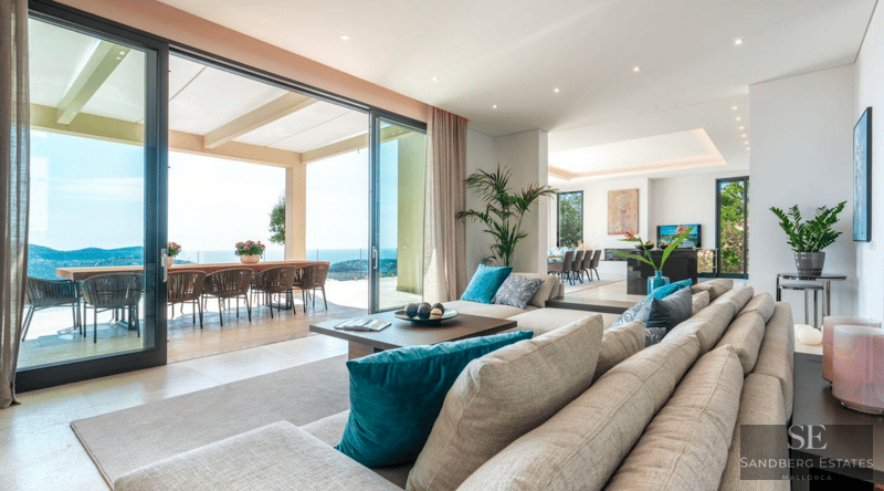 Spacious modern living room with large beige sofa, floor-to-ceiling glass doors opening to a terrace with sea views.