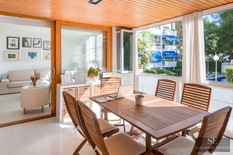 Geschlossene Holzterrasse mit Esstisch und Laptop, mit Blick auf Pool und Garten durch große Fenster.