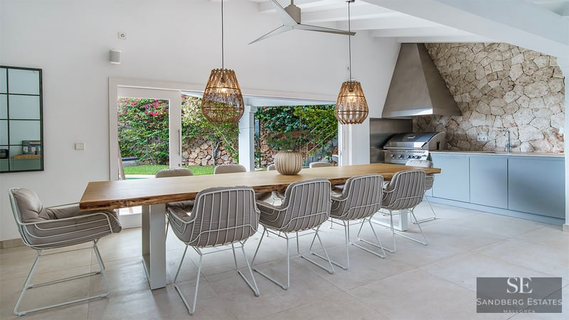 Mesa de madera maciza con sillas modernas y lámparas de mimbre junto a una cocina exterior con barbacoa de acero.