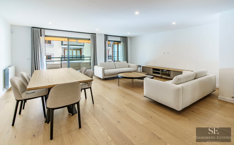 Öppen planlösning med vita soffor, matbord i trä och stora fönster som leder till en balkong.