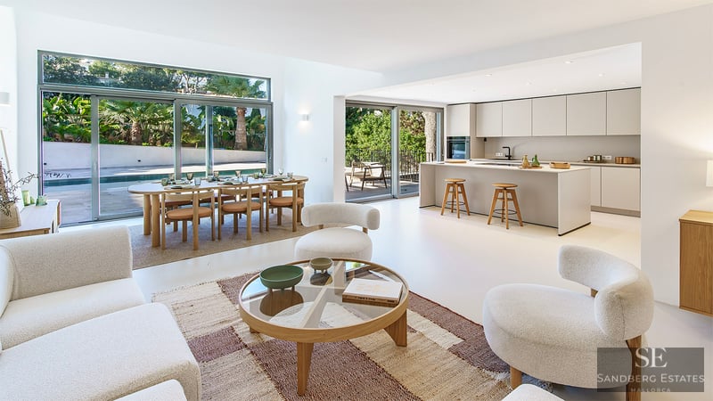 Bright open-plan room with modern white sofas, wood dining set, and minimalist kitchen overlooking a pool and garden.