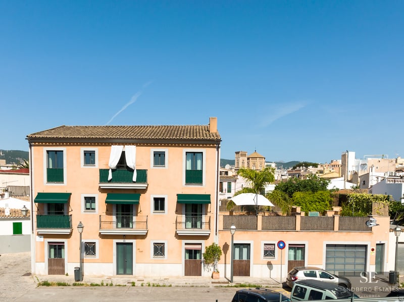 Trevånings terrakottafärgad byggnad med gröna balkonger och markiser under en klar blå himmel.