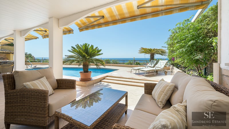 Covered terrace with wicker sofas facing a blue pool and the sea under a clear blue sky.