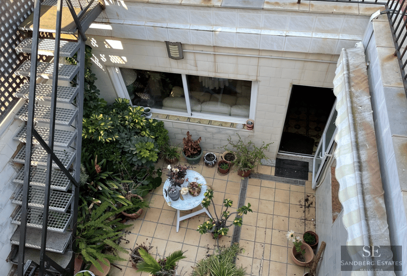 Draufsicht auf eine geflieste Terrasse mit Metalltreppe, zahlreichen Topfplanzen und einem kleinen weißen Tisch.